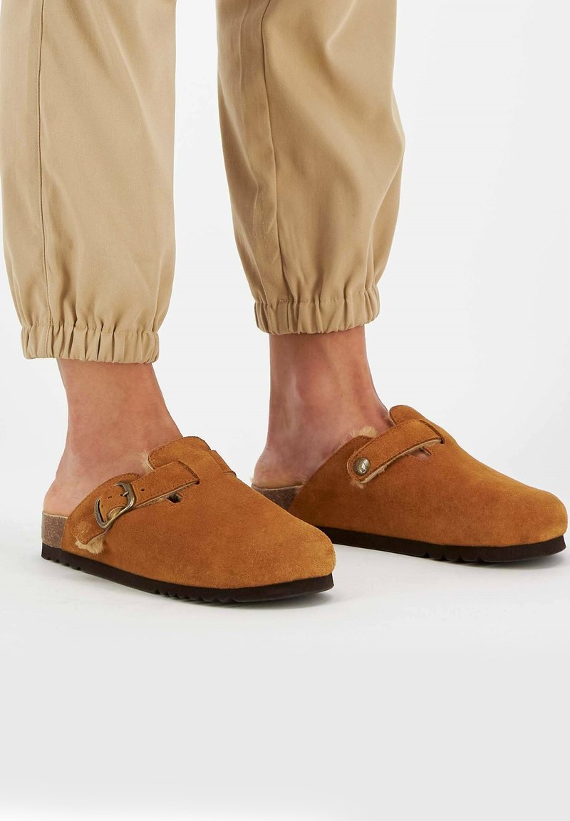 Zoccoli in suede marrone con punta rotonda, fibbia laterale e suola texturizzata, abbinati a pantaloni beige elasticizzati.