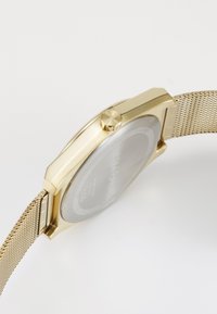 Reloj de metal dorado con correa de malla, caja rectangular y una corona prominente. La esfera presenta una parte trasera transparente con detalles de la marca.