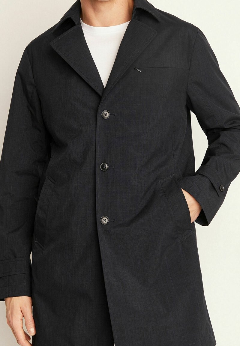 Svart, midjelång kappa av texturerat tyg med slagkrage, knappstängning, bröstficka och sidfickor.
