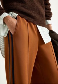 Persona che indossa un maglione marrone sopra una camicia a righe, con pantaloni marroni dotati di strisce nere ai lati, mano infilata in tasca.