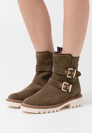 Stivaletti in suede marrone con due fibbie dorate, chiusura con cerniera e suola in gomma chiara con una trama a rilievo.