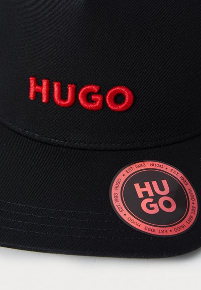 Casquette noire avec logo "HUGO" brodé en rouge à l'avant. Présente un autocollant noir circulaire avec un texte rouge indiquant l'année de création de la marque.