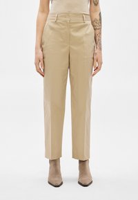 Μπεζ παντελόνι κανονικής γραμμής φορεμένο με ταμπά μυτερές μπότες αστραγάλου, το τατουάζ στο δεξί χέρι του μοντέλου είναι ορατό μπροστά σε λευκό απλό φόντο.