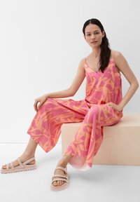 Vrouw zittend op een houten bank, gekleed in een roze en oranje gebloemde wijde jumpsuit en beige sandalen, met een witte achtergrond.