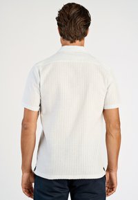 Biała koszula z krótkim rękawem o fakturowanym wzorze, z nieco dłuższym tylnym brzegiem i klasycznym kołnierzykiem. Posiada delikatne boczne rozcięcia dla większej wygody.