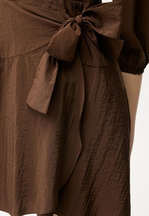 Hnědé šaty z texturované látky s velkou mašlí uvázanou v pase a krátkými nafouklými rukávy, viditelné na trupu osoby.