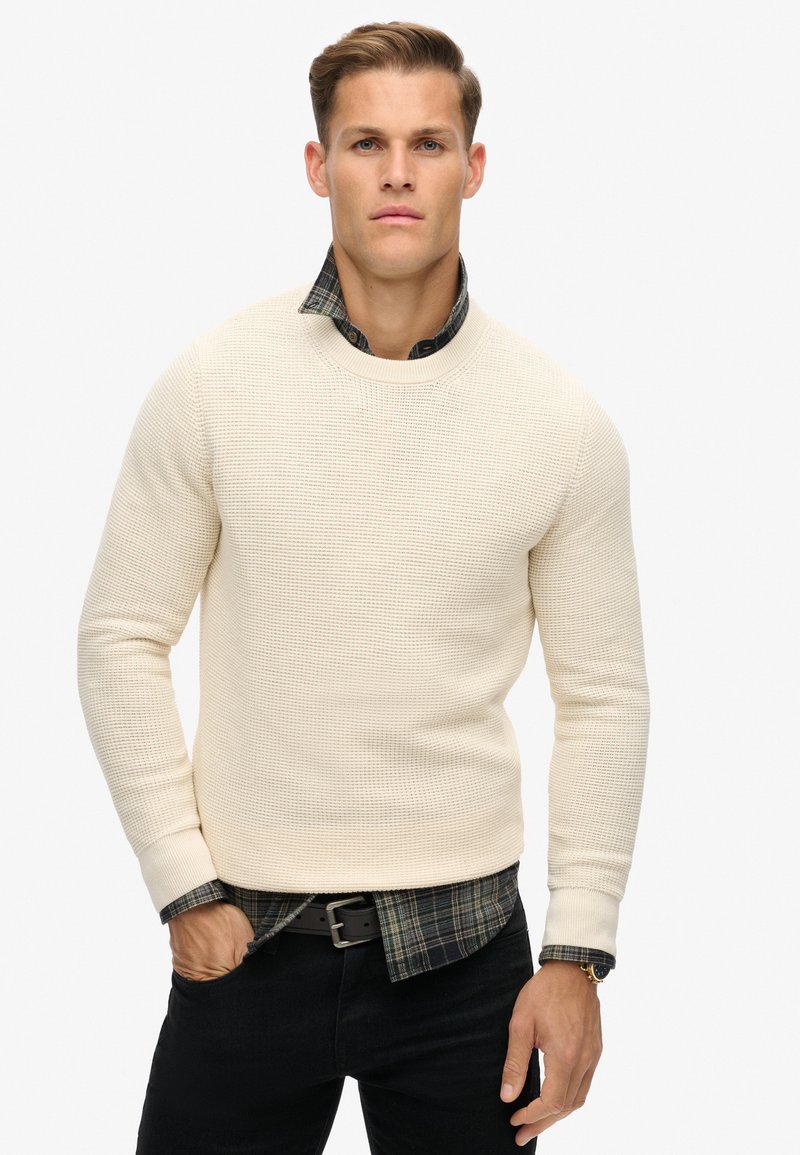 Cremefarbener, strukturiert gestrickter Pullover mit Rundhalsausschnitt, getragen über einem dunkel gemusterten Hemd und kombiniert mit schwarzen Jeans.