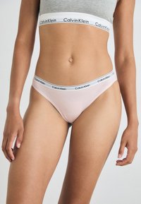 Tangá v pastelovej ružovej farbe od Calvin Klein vyrobený z jemného, elastického materiálu. Obsahuje čierno-biely pás s logom a hladkú textúru.