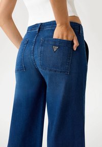 Inimene, kes kannab siniseid laia säärega teksaseid, millel on käsi tagataskus, kus on väike kolmnurkne Guess logo, ja valget ilma varrukateta pluusi.