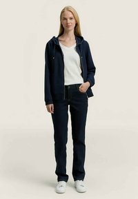 Jovem mulher de pé vestindo um hoodie azul-marinho com fecho, t-shirt branca, jeans escuros e ténis brancos contra um fundo plano.