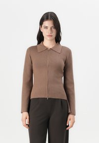 Maglione marrone con zip, texture a coste, collo piatto e maniche lunghe, abbinato a pantaloni scuri. Design semplice, con dettagli minimi.