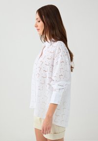 White button-up shirt made of eyelet fabric, featuring a floral pattern, long sleeves, and a relaxed fit. Paired with cream shorts.
