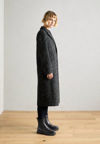 Μαύρο παλτό με στίγματα, με δομημένο γιακά, δύο κουμπιά και πλαϊνές τσέπες, συνδυασμένο με μαύρες μπότες αστραγάλου και σκούρα στενά τζιν.