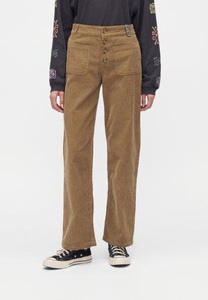 Personne portant un pantalon en velours côtelé marron taille haute avec de grandes poches avant et une fermeture par boutons, une chemise noire à manches longues et des baskets noires.