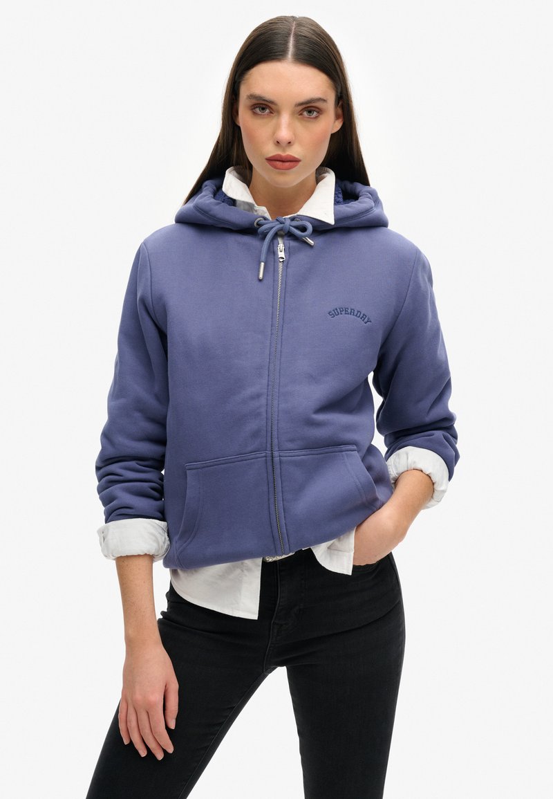 Sudadera con cremallera de color púrpura, fabricada en un tejido suave, que cuenta con una capucha ajustable, bolsillos frontales y un sutil logo en el pecho. Lleva una camiseta blanca debajo.