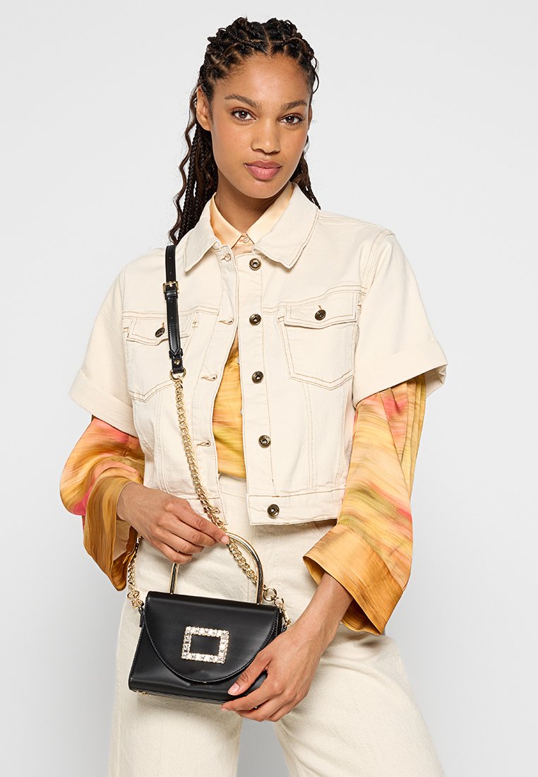 Beige, verkürzte Jeansjacke mit kurzen Ärmeln über einer bunten Batikbluse. Hält eine schwarze Handtasche mit einem mit Juwelen besetzten quadratischen Akzent.