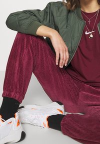 Veste bomber matelassée verte, pantalon en velours côtelé bordeaux, baskets blanches avec accents orange, colliers superposés, chaussettes noires montantes.