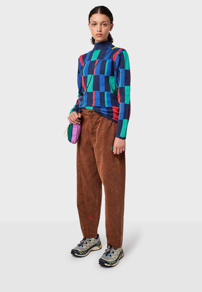 Sweter de punto a rayas multicolores con cuello alto, pantalones de pana marrones y zapatillas grises con acentos en negro y amarillo. Accesorio de bolso pequeño.