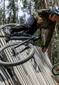 Mountain bike che naviga su una rampa di legno. La bici presenta un telaio scuro, pneumatici spessi e un sistema di sospensione. L'ambientazione include alberi sullo sfondo.