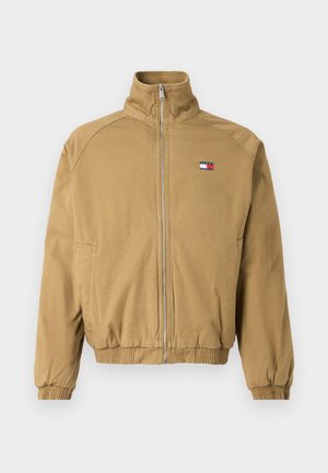 Chaqueta de color marrón claro con cremallera, cuello alto, puños elásticos y cinturilla; presenta un pequeño parche de logo con acentos en rojo, blanco y azul.