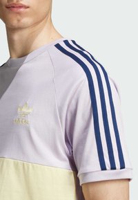 Trumpas rankovėmis lavandų spalvos marškinėliai su šviesiai geltonu apatinio skydelio. Savybės - tamsiai mėlynos juostos ant pečių ir įsiuvinėtas auksinis Adidas logotipas.