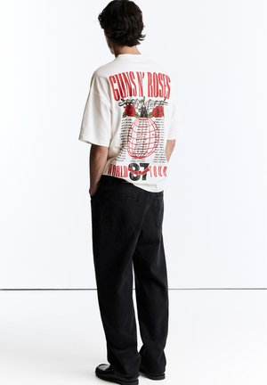 Personne portant un t-shirt blanc Guns N' Roses World Tour 87 avec des graphiques rouges et noirs dans le dos, pantalon noir, debout devant un fond blanc.