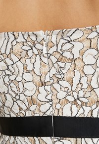 Valkoinen pitsimekko, jossa on beige pohja ja musta kukkatikkikuvio. Sisältää piilotetun vetoketjun ja leveän mustan vyötärön.