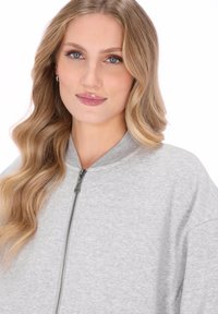 Blondynka o falujących włosach, ubrana w jasnoszary sweter na zamek, patrząca przed siebie z delikatnym uśmiechem i neutralnym makijażem.