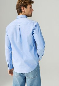 Camisa de manga larga azul claro con cuello abotonado, que presenta un pliegue en la yoke trasera y puños abotonados, combinada con unos vaqueros azul desgastado.