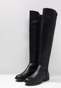 Botas largas negras de piel con punteras redondas, tacones bajos y cremalleras laterales, de pie sobre una superficie blanca con un fondo gris.