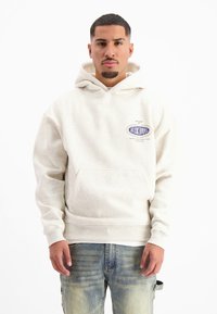 Crème-kleurige hoodie met een voorzak, met een logo op de borst. Draag het met lichtblauwe, vervaagde jeans.