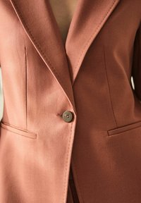Blazer v rezavé barvě vyrobený z texturované látky, s klopovými límci, dvěma předními kapsami a zapínáním na jeden knoflík.