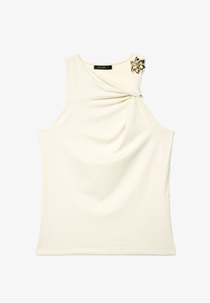 Haut crème sans manches avec un détail drapé sur la poitrine et une broche florale dorée sur l’épaule droite.