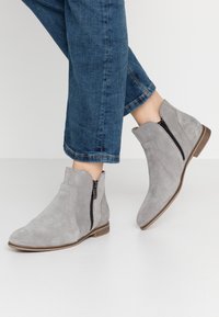 Harmaat mokkasiinit nilkkasaappaat, joissa on sivuvetoketju ja kumipohja. Saappaissa on sileitä ja teksturoiduja yksityiskohtia varressa.