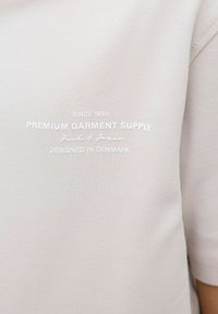 Helebeežist särk, millel on tekst "Premium Garment Supply" koos märksõnaga "Kujundatud Taanis", rõhutades brändi pärandit.