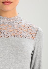 Grå långärmad topp med blommigt spetsdetalj på axeln och en vågig halsringning. Slät bomullsblandning, figurnära design.