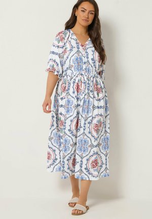 Kobieta ubrana w białą sukienkę midi z niebiesko-różowymi kwiatowymi wzorami i bufkami na rękawach, w połączeniu z beżowymi, plecionymi sandałami na płaskim obcasie.