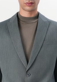 Grå skräddarsydd kavaj med texturerad yta, enkelknapp, flikiga slag, över en åtsittande brun polotröja. Skarp, modern design.