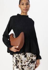 Brown faux leather bag with a curved shape, held in a hand. Model wears a black, textured, ruffled blouse and a patterned, flowy skirt.