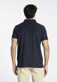 Polo shirt navy en coton avec des manches courtes et un col. Doté d'une coupe droite et d'un petit logo accent près de l'ourlet. Fond neutre.