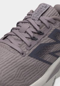 Κοντινό πλάνο γκρι αθλητικού παπουτσιού με υφασμάτινη υφή, κορδόνια, σκοτεινό λογότυπο και τη λέξη "FuelCell Propel" στη γλώσσα και στη σόλα.