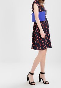 Merekkoppa tummansininen mekko punaisella kukkakuviolla, yhdistettynä siniseen clutch-laukkuun ja mustiin nilkkasaappaisiin. Mekossa on lyhyet hihat ja liehuva muotoilu.