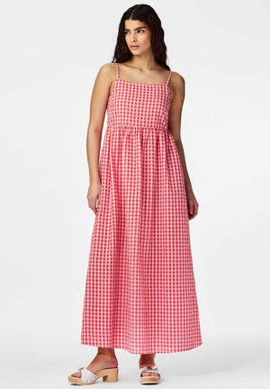Ženska z dolgimi temnimi lasmi, oblečena v rdečo-belo karo brezrokavno maxi obleko in bele sandale z visokimi petami, stoji in gleda levo.