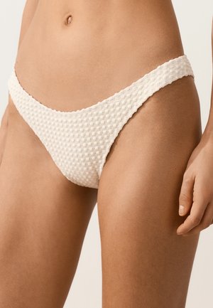 Onderlichaam van een persoon die een crèmekleurige, gestructureerde bikinibroek draagt met een hoog uitgesneden ontwerp tegen een effen achtergrond.