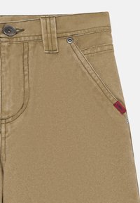 Beige katoenen broek met een gladde textuur, voorzien van een metalen knoop, een metalen rivet en een klein rood label dat in de tailleband is gestikt.