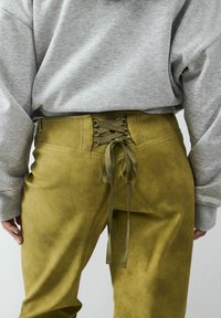 Zielono-oliwkowe spodnie z sztucznego zamszu o wysokim stanie, z zapięciem na sznurówki z tyłu, ozdobione dopasowaną wstążką. Miękka faktura i minimalne detale szwów.