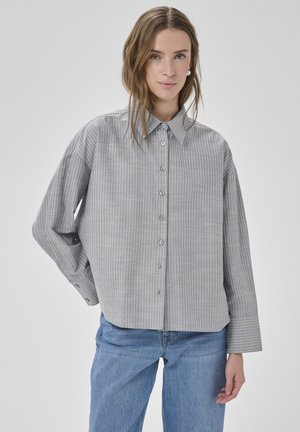 Kobieta ubrana w luźną, szarą koszulę w prążki z szerokimi mankietami oraz jasnoniebieskie dżinsy, stojąca na tle jednolitego, szarego tła.