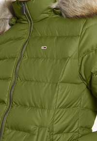 Olivengrøn, polstret jakke med høj krave, pelskant og lynlås. Har kviltet struktur og et lille logo på brystet.