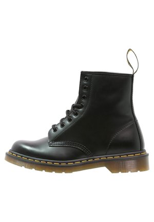 Bottine en cuir noir avec huit œillets, semelle épaisse translucide, coutures jaunes et boucle de traction à l'arrière.