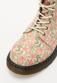 Botte avec base beige, décorée de motifs floraux roses et verts, lacets crème, semelle noire avec couture jaune et semelle marron.
